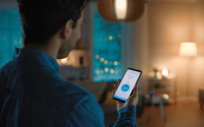 Young Handsome Man Gives a Voice Command to a Smart Home Application on His Smartphone To Switch on the Lights and Set a Comfortable Temperature. It's a Cozy Evening in Living Room.
