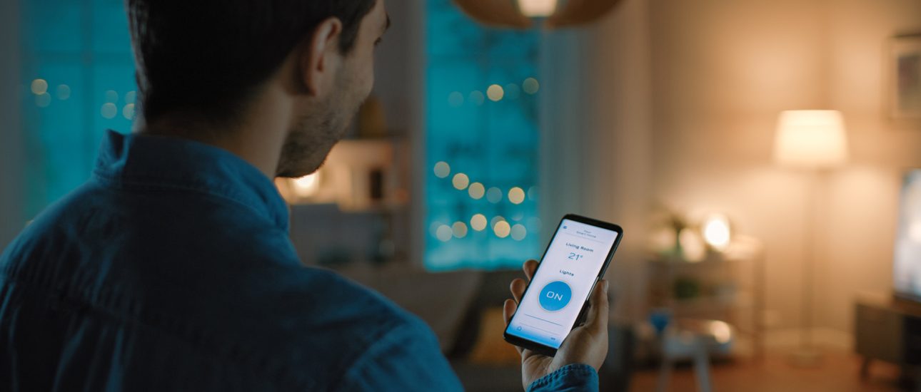 Young Handsome Man Gives a Voice Command to a Smart Home Application on His Smartphone To Switch on the Lights and Set a Comfortable Temperature. It's a Cozy Evening in Living Room.